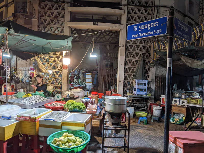 Night market stall with baskets of produce, seafood, and herbs on display, illuminated by hanging lights on Preah Chanreachea Street.