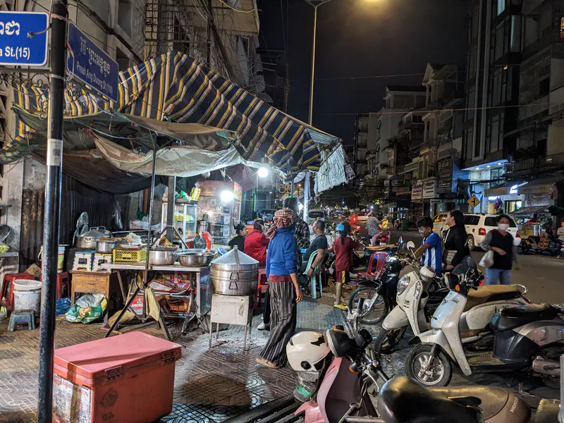 Street food cart at night with skewers of meat, dried fish hanging, and fresh vegetables on display while two men stand nearby.