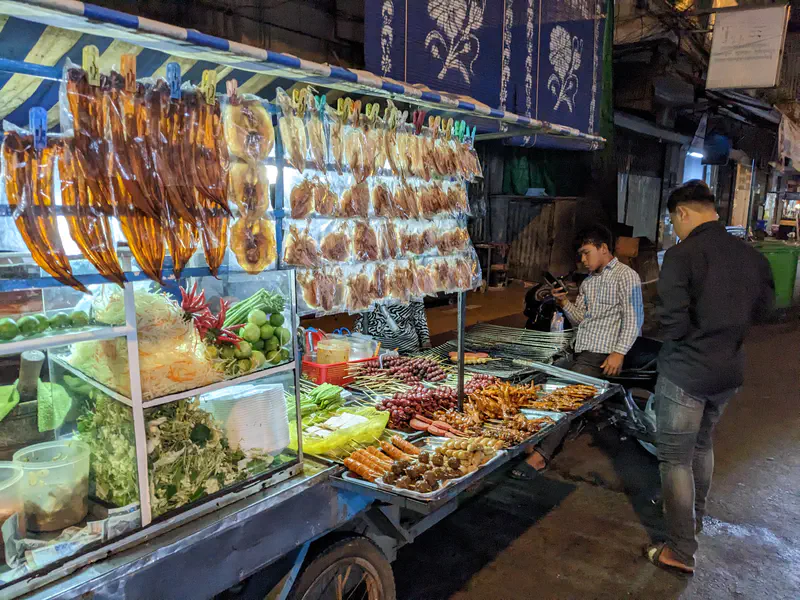 Nighttime street scene with food stalls, steaming pots, and people gathered around scooters and tables under bright lights.
