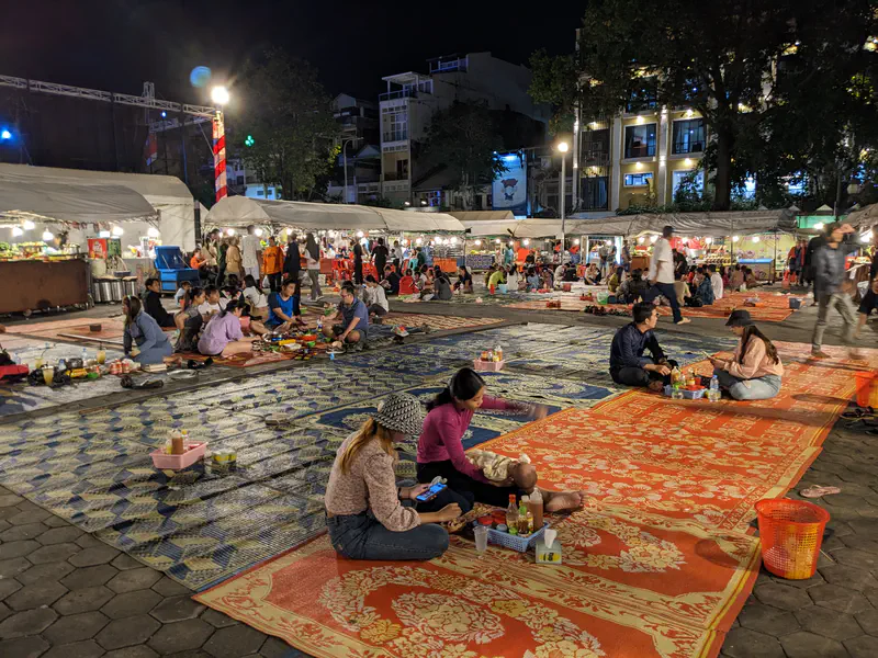 People sitting on patterned mats eating at a lively night market with food stalls in the background.