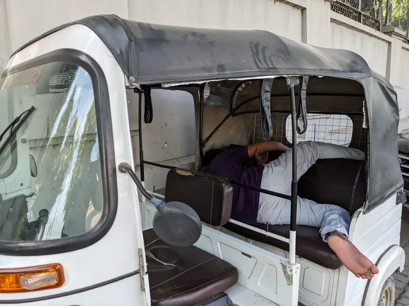 Person lying down and resting inside a parked tuk-tuk on the side of the street.