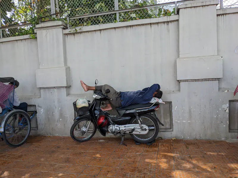 Man lying on a parked motorcycle by a wall, taking a nap in the shade.