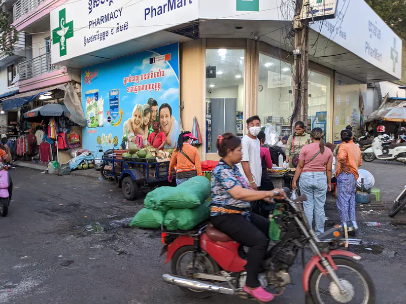 Street scene with people gathered outside a pharmacy and a woman riding a motorbike with green sacks.