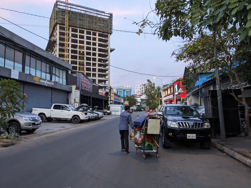 Person pushing a cart with bags along a city street lined with parked cars and a tall building under construction.