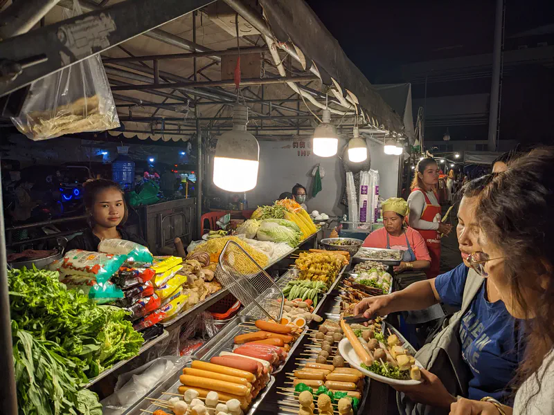 People selecting skewers and vegetables from a busy night market food stall.