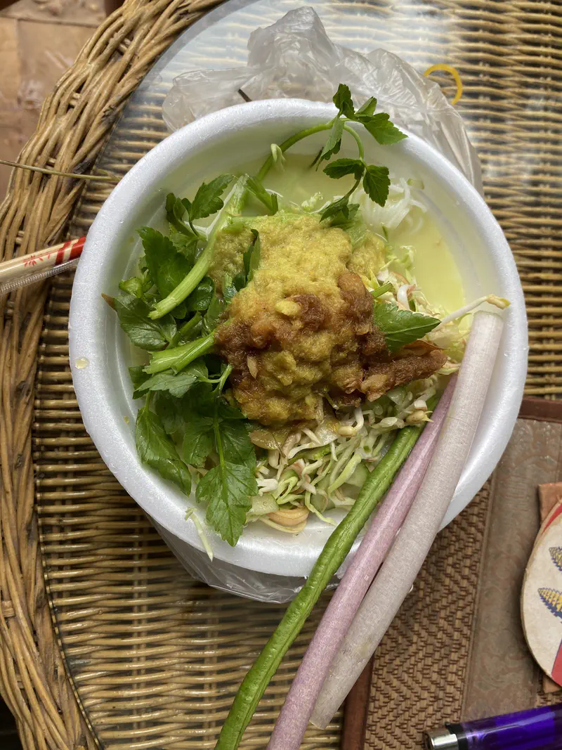 Bowl of Cambodian noodle soup with fresh herbs, long beans, sprouts, and a yellow curry sauce served in a foam bowl.