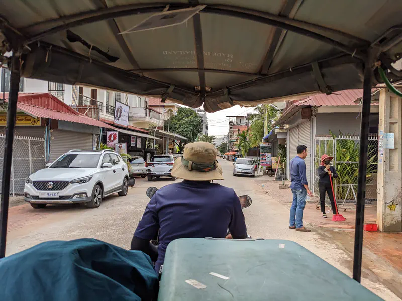View from inside a tuk-tuk showing the driver in a hat navigating a narrow street with parked cars and people sweeping the sidewalk.