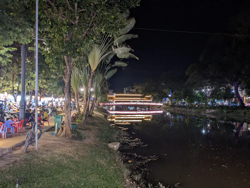 Night view of a riverside with trees, parked motorbikes, and a bridge decorated with glowing lights reflecting in the water.