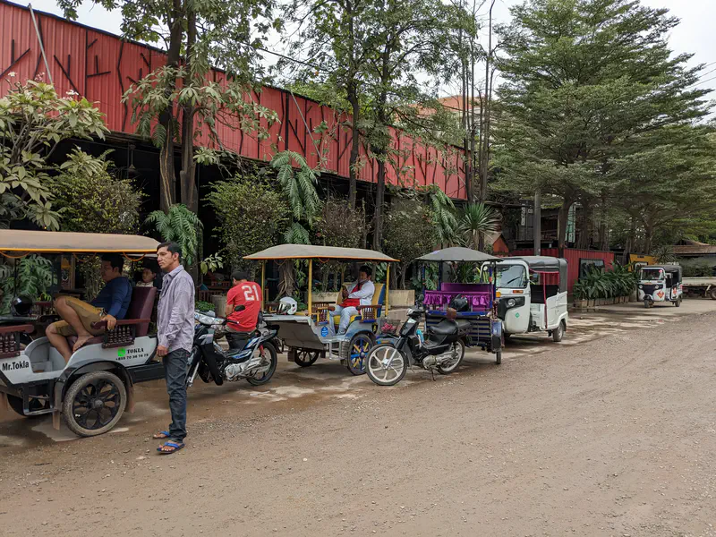 Row of tuk-tuks parked along a dirt road outside a red building, with drivers waiting nearby under trees.