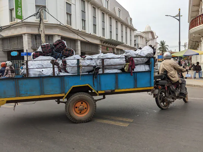Motorcycle pulling a large blue trailer loaded with sacks and bundles, driving on a city street near white buildings.