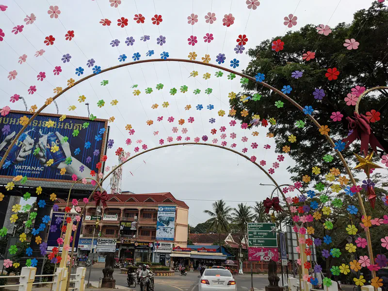 Street decorated with colorful hanging flower ornaments on arches, with traffic and buildings in the background during the day.
