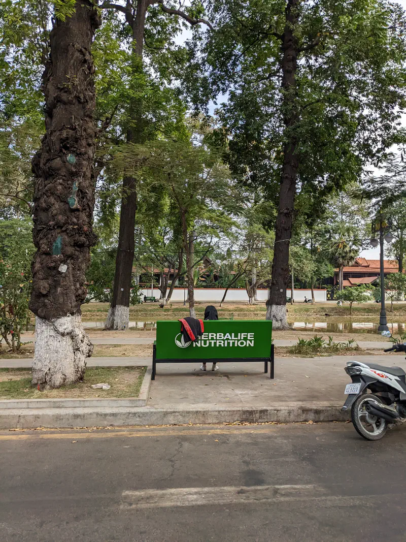 Green park bench with Herbalife Nutrition logo, a person sitting with their back facing, and trees in the background.