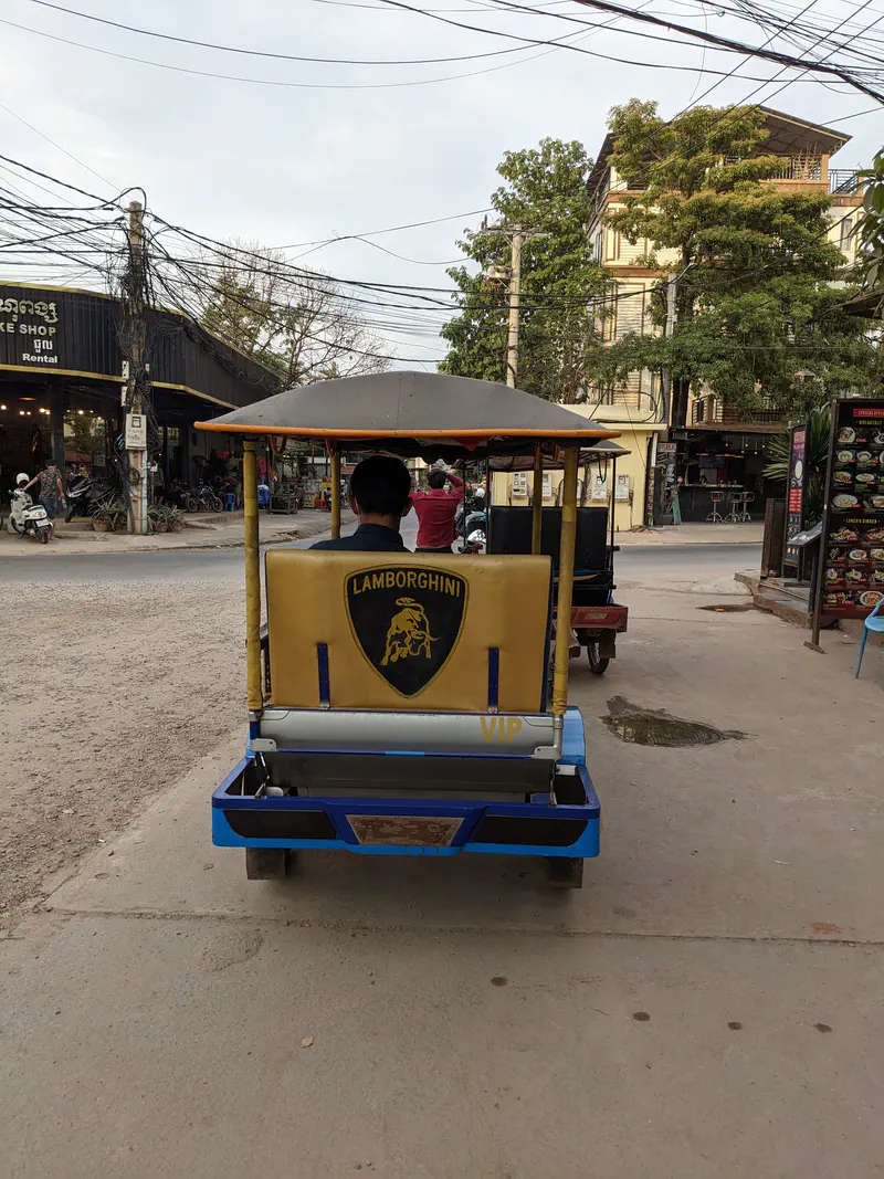Rear view of a tuk-tuk with a Lamborghini logo on its backrest, parked on a street with shops and power lines visible.