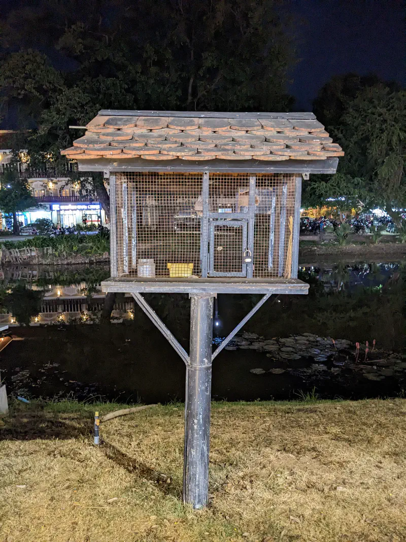 Small wooden cage structure on a tall pole by a riverbank at night, with a tiled roof and mesh walls.