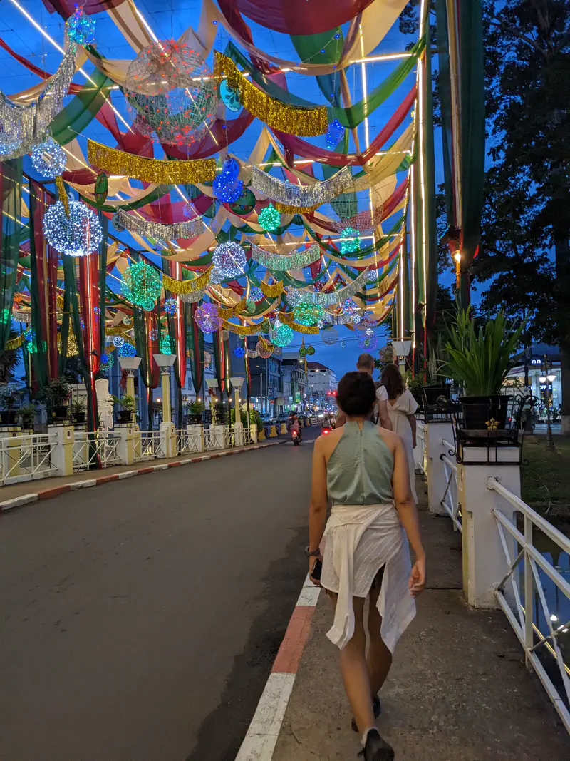 Street at night with festive overhead decorations of lights, ribbons, and glowing ornaments, with people walking along the sidewalk.
