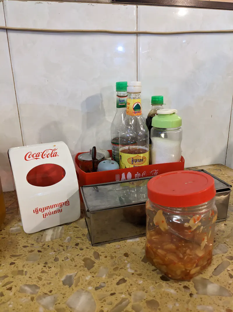 Restaurant table with condiments including sauces, sugar, pickled garlic in a jar, and a Coca-Cola napkin holder.