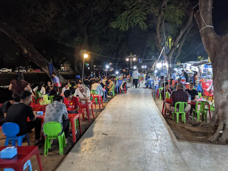 Busy Cambodian night market with people sitting on colorful plastic chairs at small tables under trees, eating and socializing along a pedestrian walkway.
