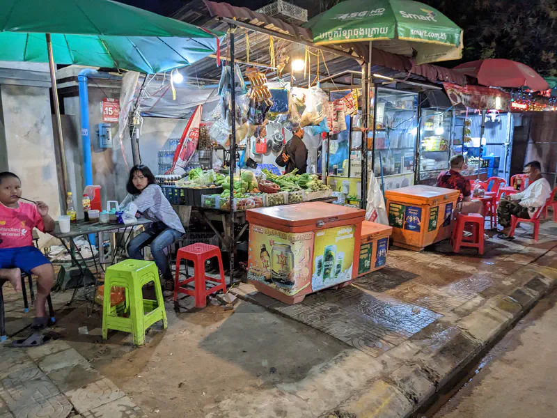 Street food stall at night with plastic stools, coolers, and fresh produce on display, with people dining nearby.