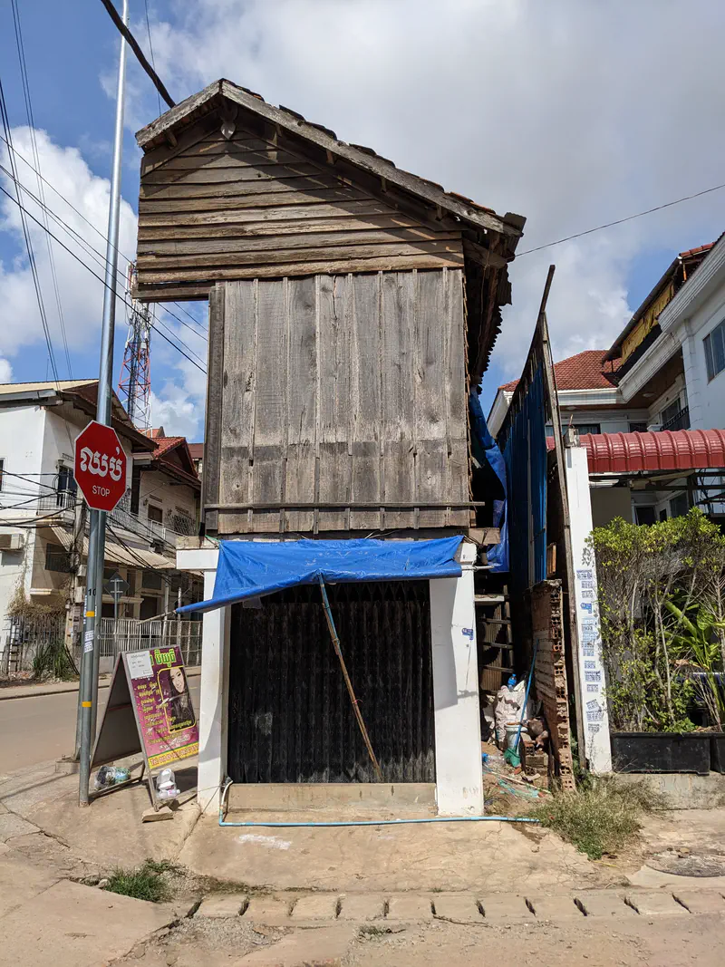 Old wooden house in Cambodia with weathered planks, a corrugated roof, and a blue tarp shading the lower entrance, situated beside newer concrete buildings.