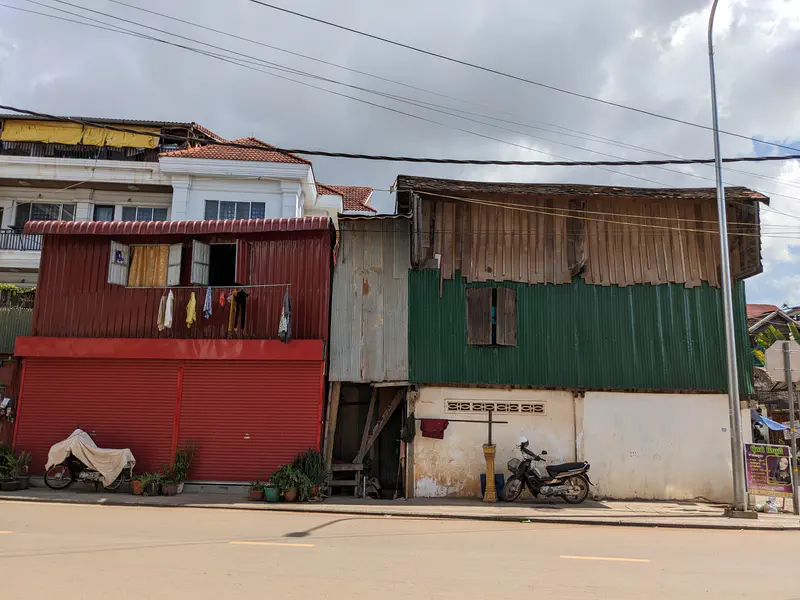 Row of old Cambodian shophouses with corrugated metal walls, one painted red with clothes hanging outside and another with green siding, with motorbikes parked below.