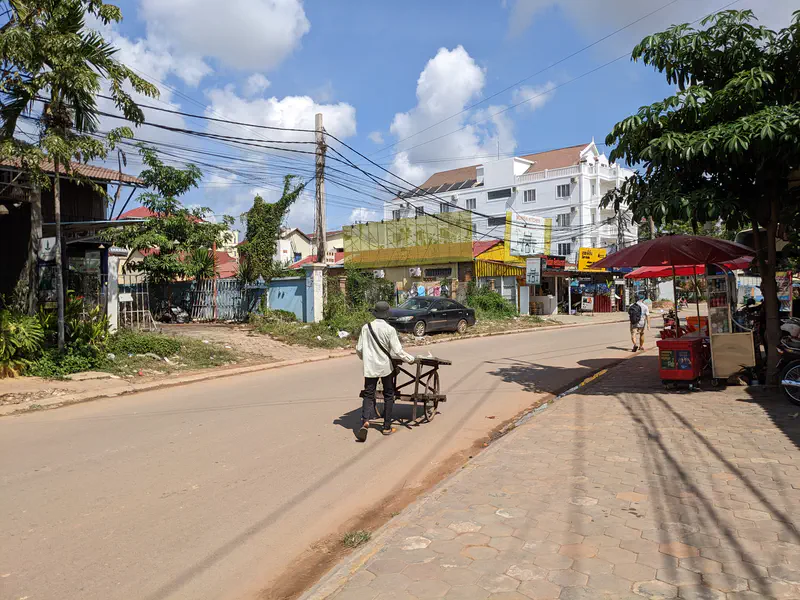 Man pushing a wooden cart along a sunny street in Cambodia, with small shops, a street vendor under a red umbrella, and white buildings in the background.