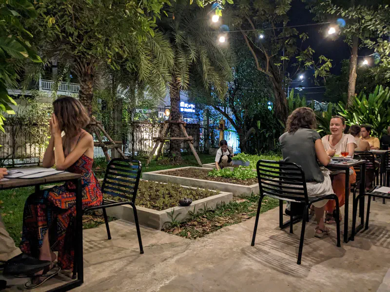 Outdoor garden restaurant at night with diners sitting at black metal tables, string lights overhead, and a person tending to plants in the background.