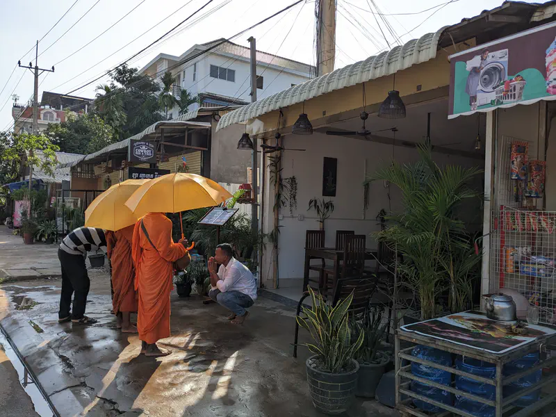 Two Buddhist monks in orange robes holding yellow umbrellas receive offerings from locals outside a small shop in Cambodia.