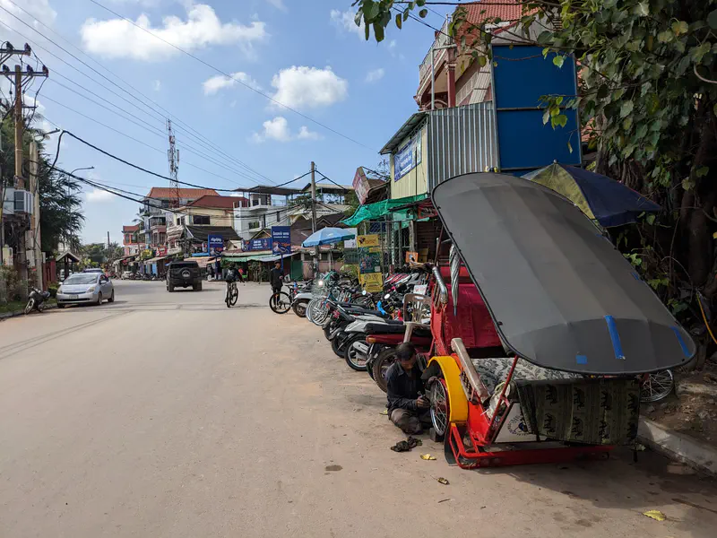 Street scene with motorcycles parked along the side and a man repairing a rickshaw under its large canopy.