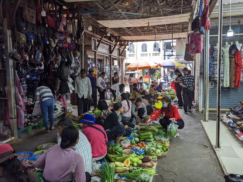 Busy Cambodian market scene with vendors sitting on the ground selling fresh vegetables, fruits, and herbs, while shoppers browse stalls selling clothes, bags, and accessories.