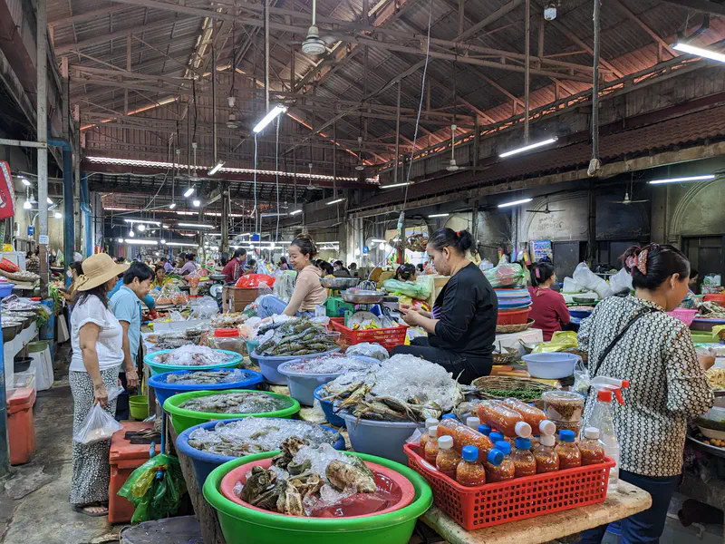 Cambodian wet market with vendors selling fresh fish, crabs, and seafood displayed on ice in large plastic tubs, alongside bottles of sauces and condiments, under a high wooden roof.