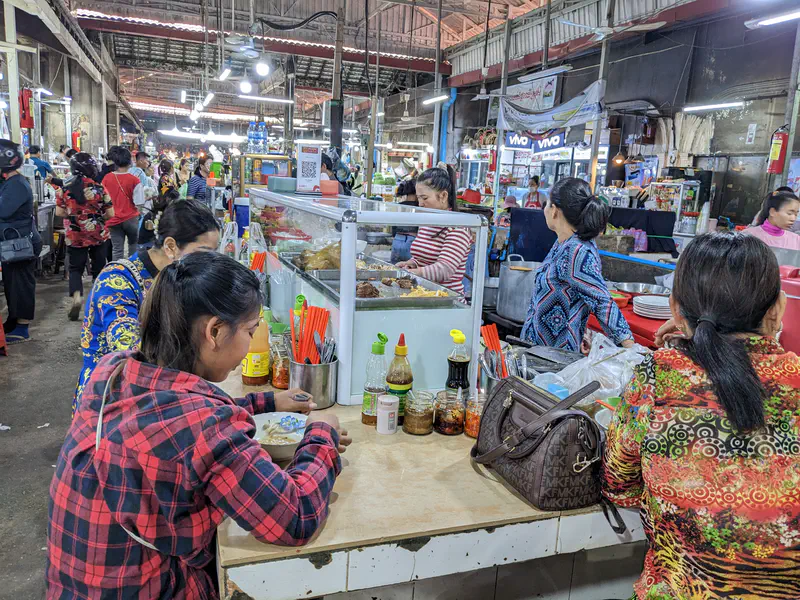 Busy Cambodian market food court with women sitting at a counter eating noodles, condiments and chopsticks on the table, and food vendors preparing dishes behind glass displays.