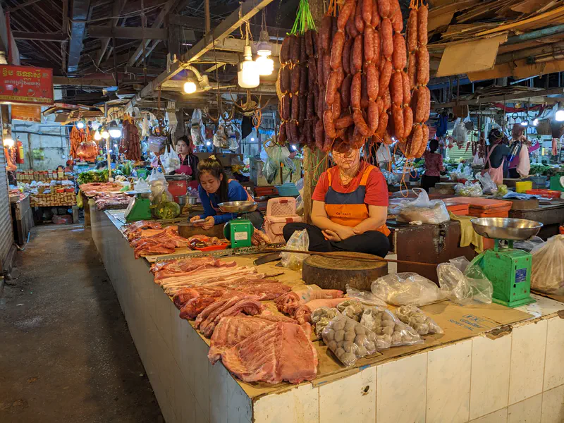 Butchers at a Cambodian market selling fresh cuts of pork, sausages, and other meats displayed on tiled counters, with hanging lights overhead and vendors working behind the stalls.