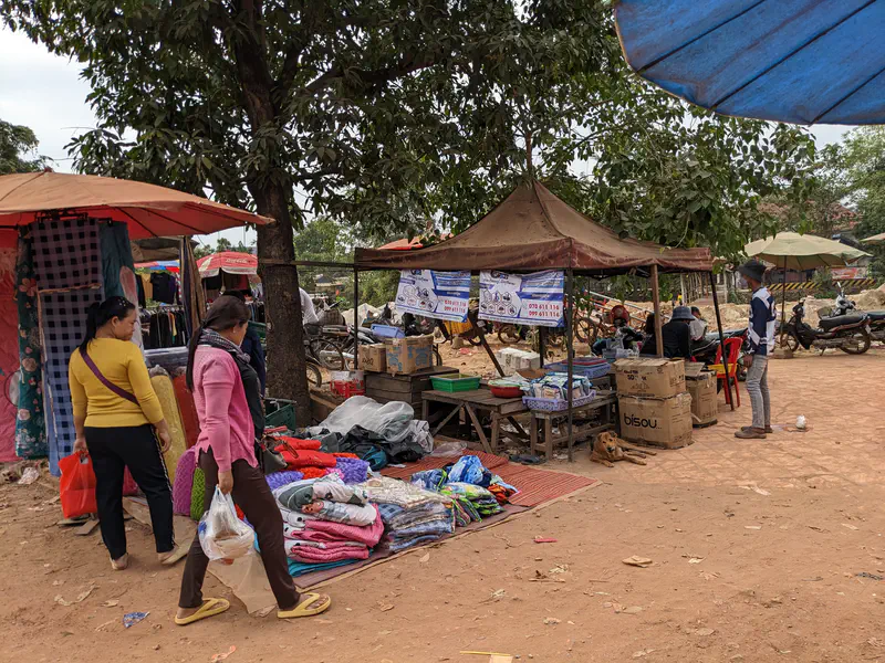 Outdoor market scene with women walking past folded blankets and fabrics displayed on the ground, vendors sitting under makeshift tents, and a dog resting in the shade among cardboard boxes.