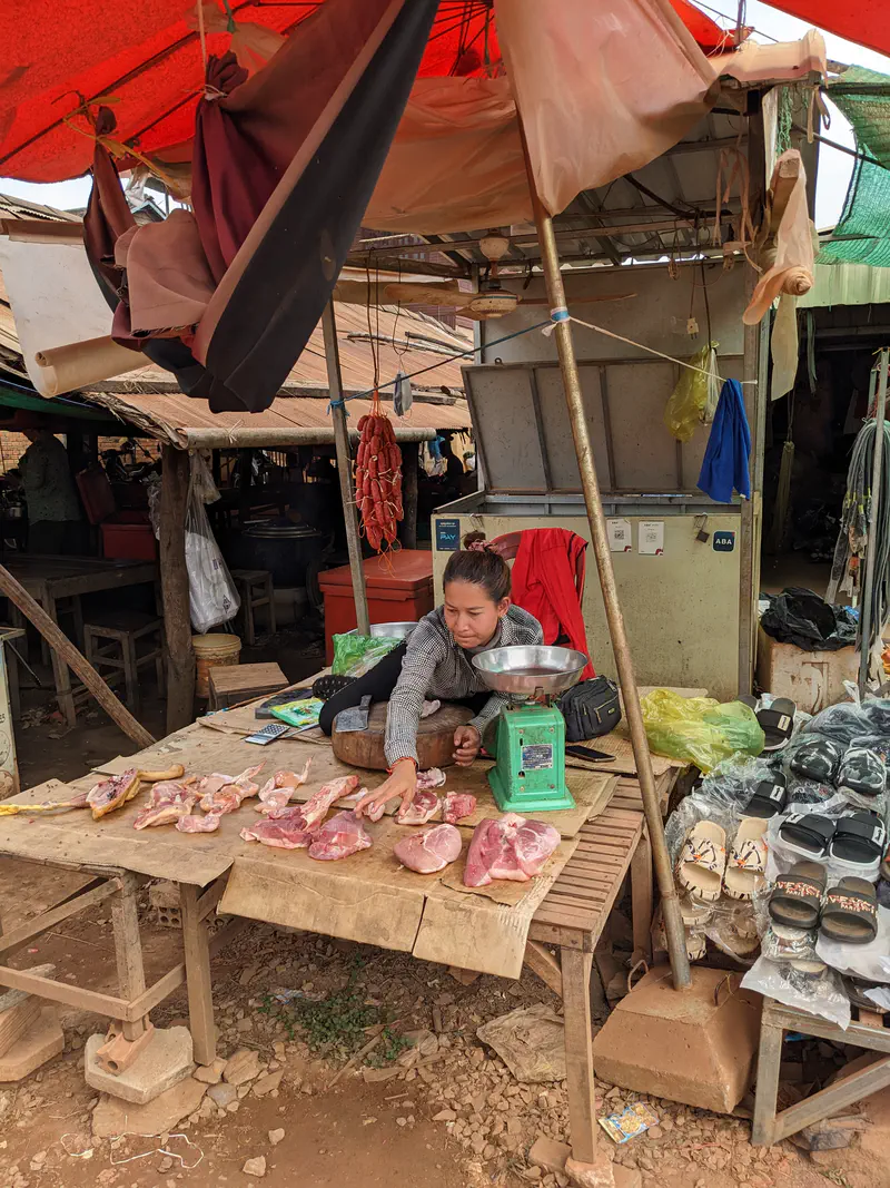 Woman at an open-air market stall lying on a wooden table as she arranges pieces of raw meat next to a green weighing scale, with sandals and other goods displayed nearby.