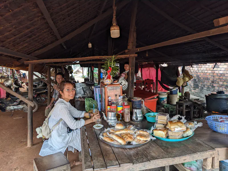 Rustic roadside eatery with wooden tables covered in bread rolls and packaged pastries, where a woman with glasses sits smiling with a drink, surrounded by locals in a shaded hut.