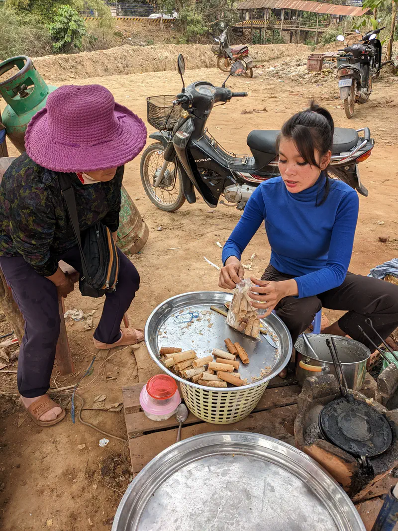 Street vendor in a blue sweater sitting beside a large tray of rolled wafer snacks, placing them into a plastic bag for a customer in a purple hat, with motorbikes parked in the background.