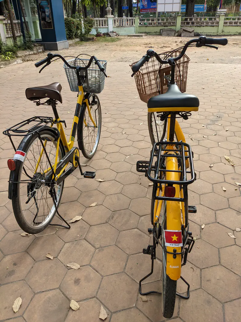Two yellow rental bicycles with baskets parked on a tiled pavement, one displaying a Vietnam flag sticker on the rear fender.
