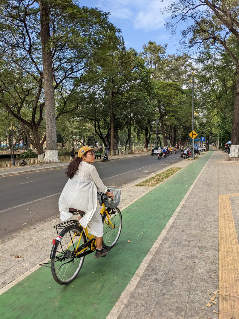 Person riding a yellow rental bicycle with a basket along a green bike lane next to a tree-lined street with cars and motorbikes.
