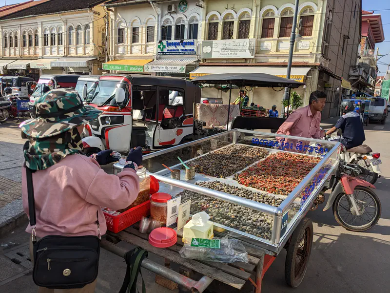 Street vendor in Cambodia selling snails and shellfish from a glass display cart attached to a motorcycle, with tuk-tuks and colonial-style buildings in the background.