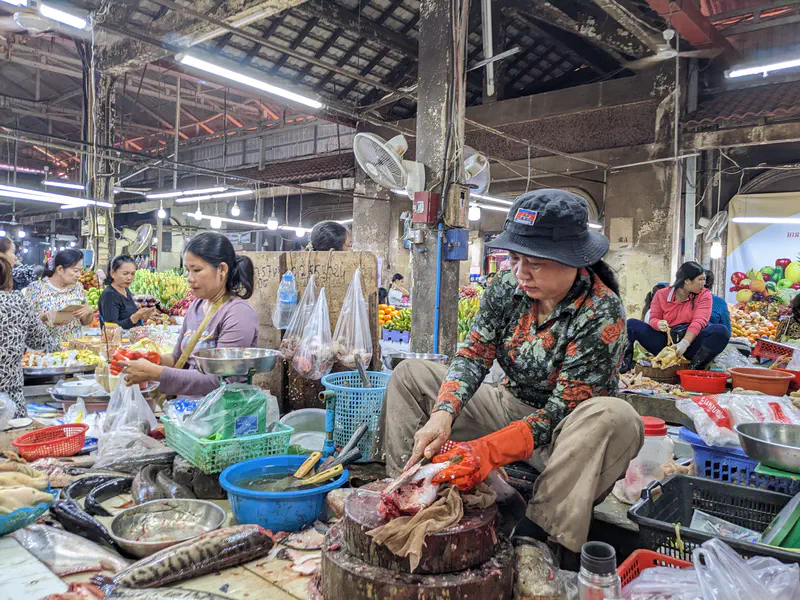 Fish vendor at a Cambodian market wearing a hat and gloves while cutting fresh fish on a wooden block, surrounded by baskets of seafood, vegetables, and other sellers in the busy marketplace.