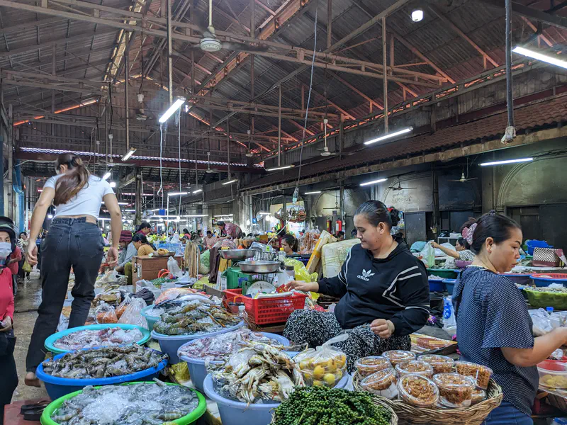 Vendors at a Cambodian wet market selling fresh seafood including shrimp, crabs, and fish on ice in large tubs, alongside baskets of green pepper and jars of condiments, under a wooden roof with bright lights.