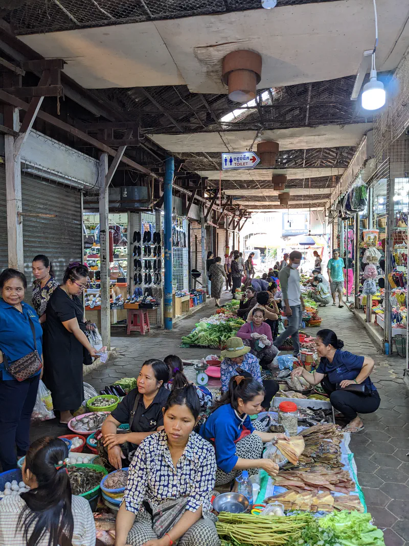 Vendors at a Cambodian market sitting on the ground with baskets of vegetables, dried fish, eggs, and fresh produce, while shoppers browse through nearby stalls selling clothes and shoes.