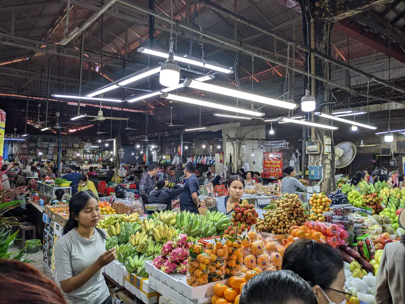 Indoor Cambodian market with vendors selling colorful fresh fruits such as bananas, oranges, dragon fruit, grapes, and longan under fluorescent lights.