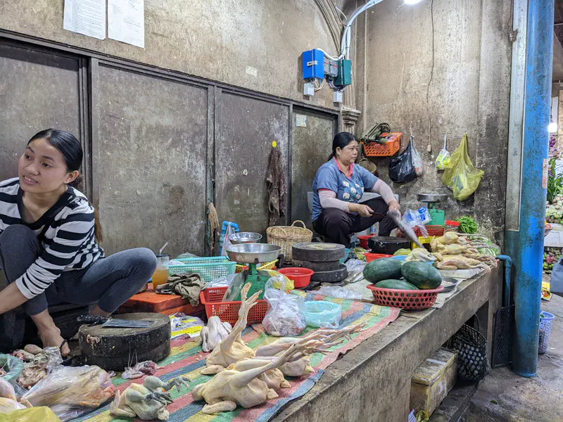 Two women at a Cambodian market poultry stall displaying whole plucked chickens, chicken parts, and some vegetables on mats, with butchers preparing meat for customers.