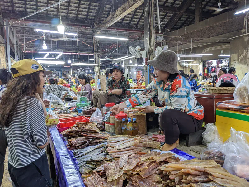 Vendor at a Cambodian market stall selling dried fish and seafood, handing goods to a customer, with jars of condiments and bottles of sauce on the counter in a busy market setting.