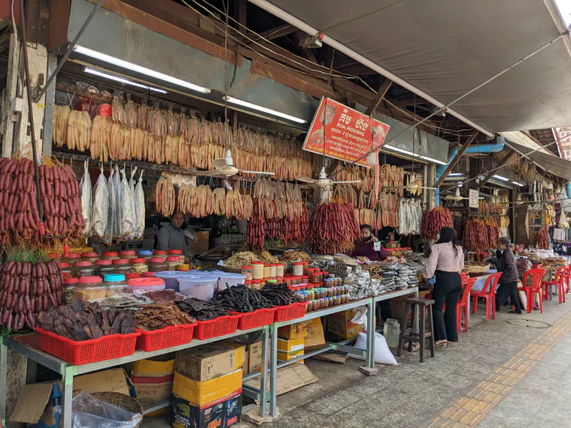 Traditional Cambodian market stall displaying dried meats, sausages, and fish hanging overhead and arranged in baskets and jars on tables, with vendors attending to customers.