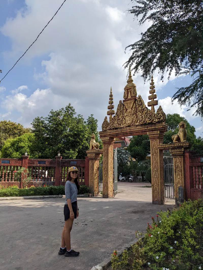 Woman standing in front of an ornate golden temple gate with intricate carvings and statues of lions, surrounded by greenery under a partly cloudy sky.