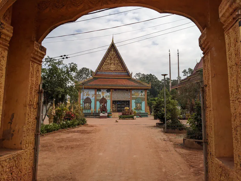 View through an ornate archway into a temple courtyard with a richly decorated building featuring golden carvings and colorful murals, surrounded by plants and statues.