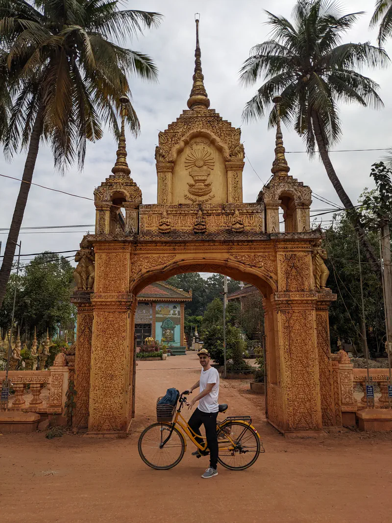 Man standing with a yellow bicycle in front of an ornate golden temple gate decorated with carvings and spires, flanked by tall palm trees on a dirt path.