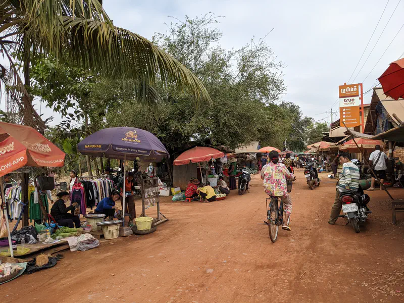 Bustling village market on a dirt road with stalls selling clothes and produce under umbrellas, people walking, motorcycles parked along the sides, and a person cycling down the center.